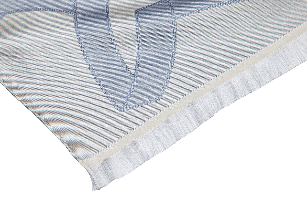 A close-up of a white woven textile with light blue stitched geometric lettering and white fringe along one edge, shown on a white background.