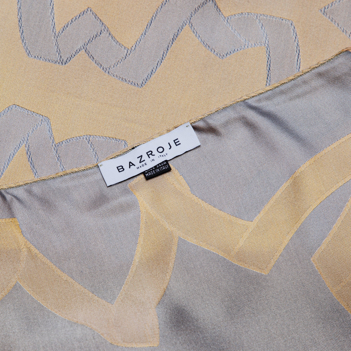 A close-up of a BAZROJE brand scarf featuring a geometric pattern in beige and gray, with a white brand label sewn onto the edge.