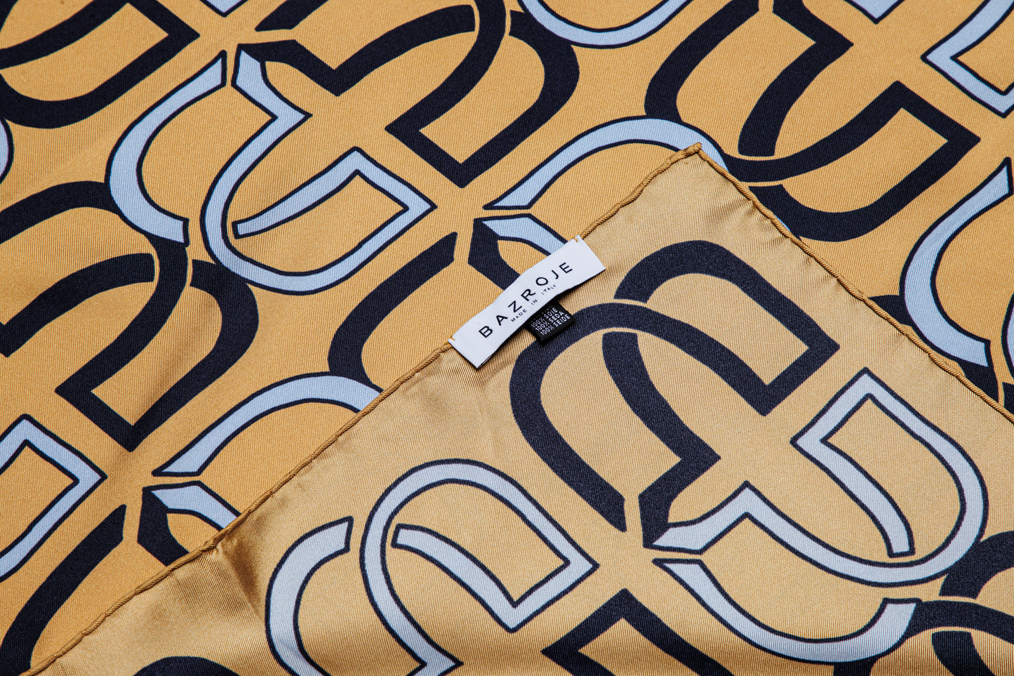A close-up of a patterned scarf or fabric with a repeating interlocking monogram design in black, navy, and light blue on a warm tan background. A small white fabric tag with the brand name 'BAZROJE' is visible near the center.