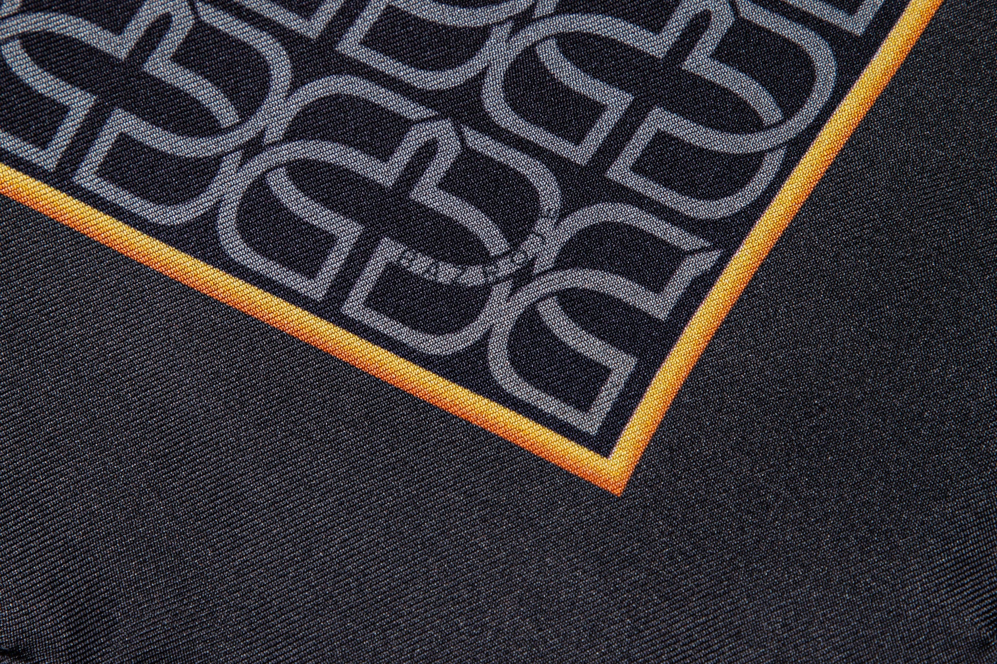 Close-up of a black scarf featuring a repeating gray interlocking monogram pattern and a bright orange border.