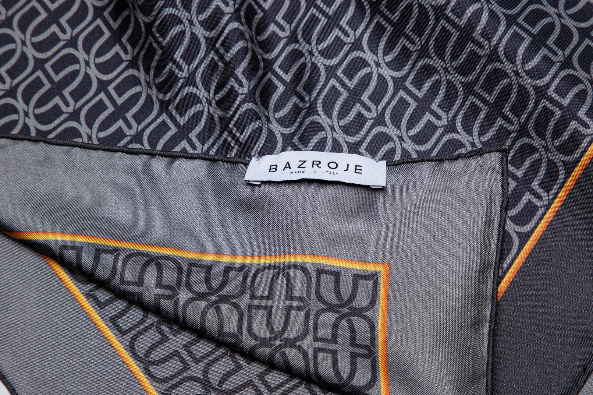 Close-up of a patterned scarf or fabric with a white BAZROJE label, featuring a dark gray and black interlocking geometric pattern with a thin orange border on a gray satin-like surface.