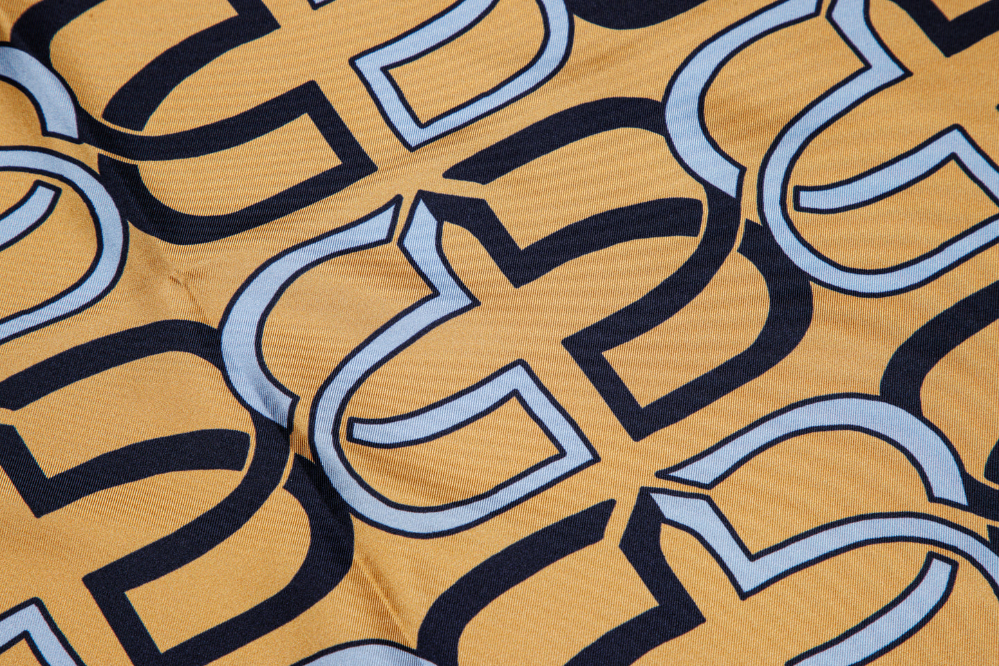 A close-up of a fabric or material surface with an allover repeating interlocking geometric pattern in mustard yellow, dark navy, and light blue.